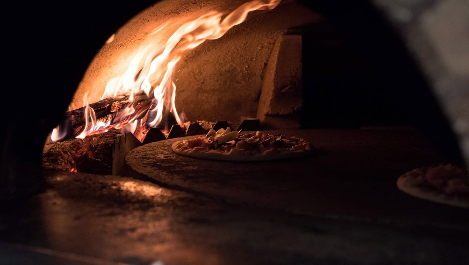 L'intérieur du four à bois : les bûches en flammes au fond, et une pizza qui cuit sur la pierre, à l'entrée