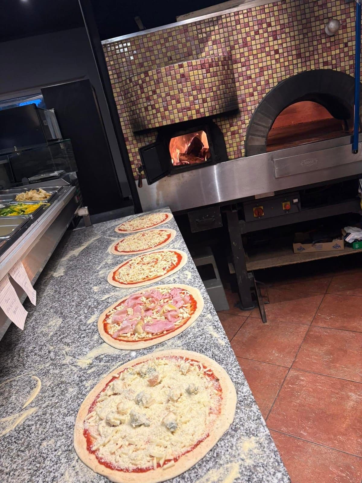 Le four à bois habillé de mosaïque, allumé, et devant lui six pizzas crues alignées sur le plan de travail en granit